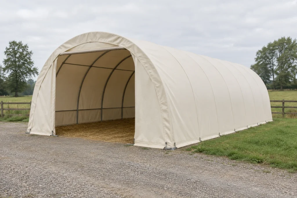 Illustration d’un abri tunnel clair doté d’un toit en arc, d’une entrée ouverte et d’un sol couvert de paille, près d’une prairie clôturée.